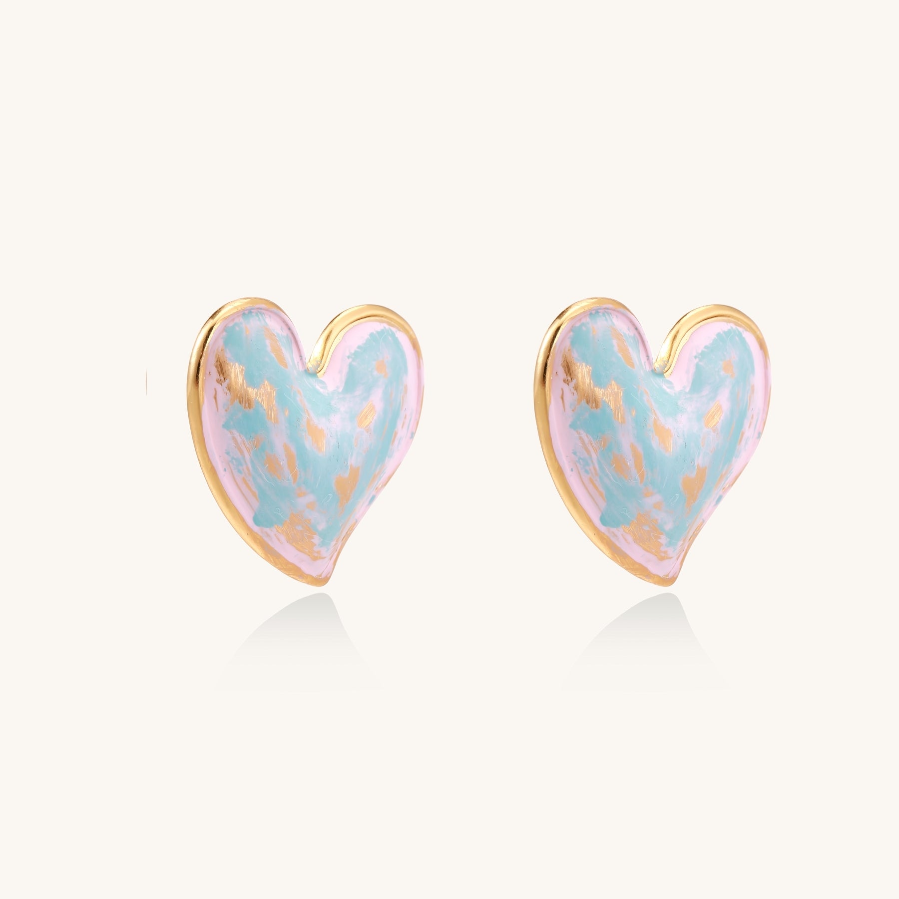 Heart-shaped stud earrings with iridescent blue-pink enamel finish and gold-plated brass frames against cream background