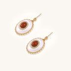 Gold-plated drop earrings with oval mother-of-pearl stones, central red gemstones, and crystal accents on white background.